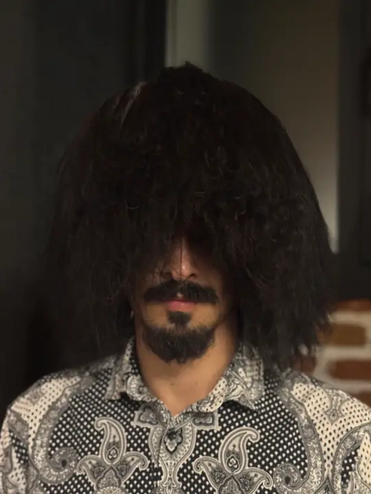 Black sheepskin papakha with long wavy wool covering the eyes, worn by a man in a patterned shirt, front view