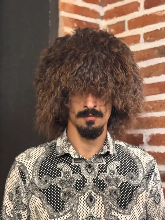 Goat fur papakha with long curly light brown fur, worn by a man in a patterned shirt, front view, brick wall behind