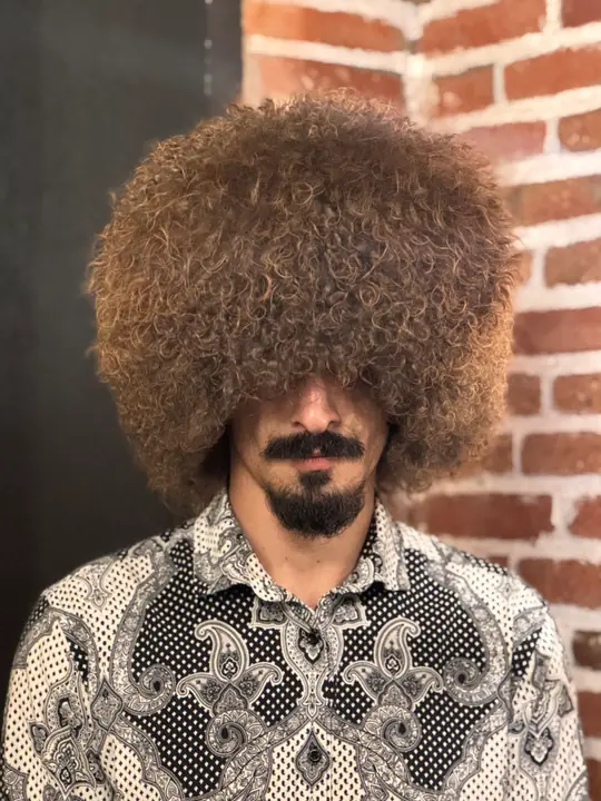 Goat fur papakha with dense curly brown fur, worn by a man in a patterned shirt, front view, brick wall behind