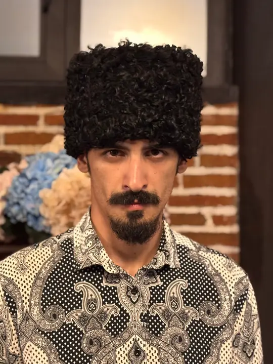 Black karakul papakha with short tight curls, worn by a man in a patterned shirt, front view