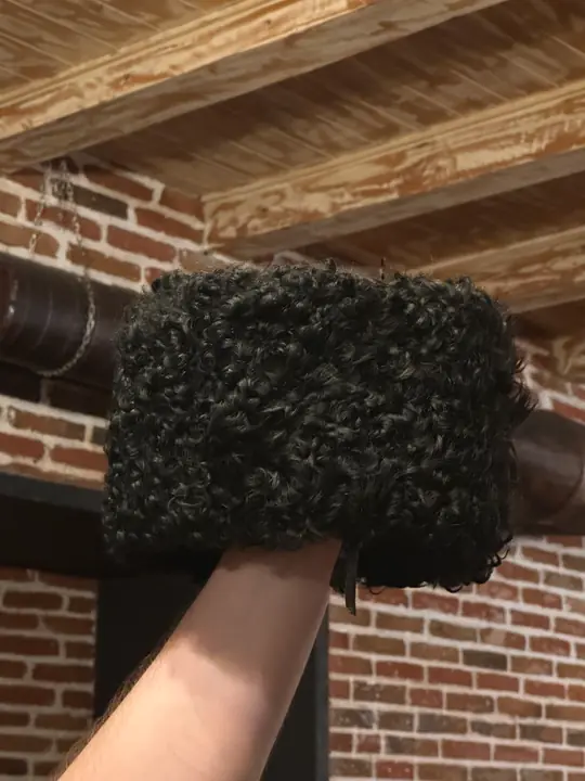 Black karakul papakha held up in a hand, seen from the side, wooden ceiling behind
