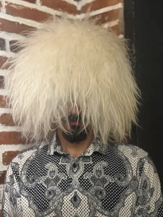 White sheepskin papakha with long straight wool, worn by a man in a patterned shirt, front view, brick wall behind