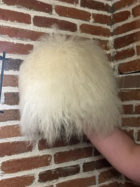 White sheepskin papakha held in a hand against a brick wall, seen from the side