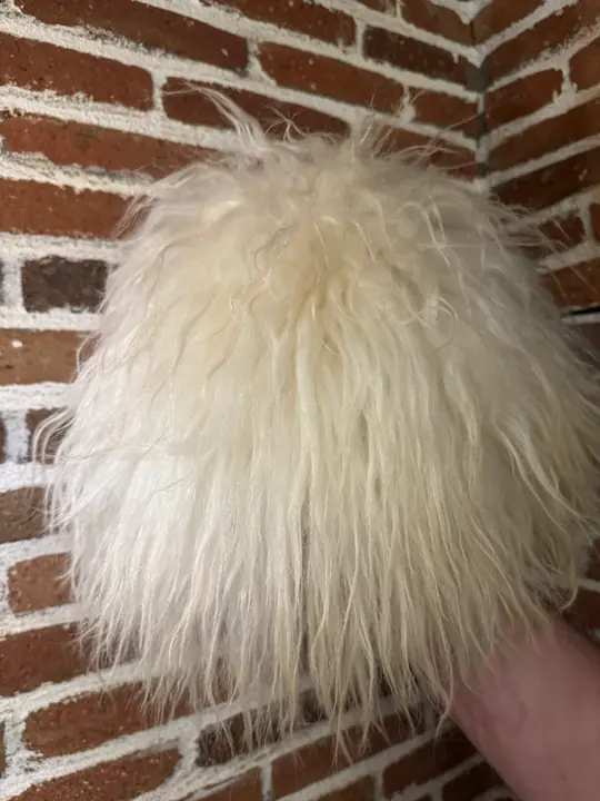 White sheepskin papakha held in a hand against a brick wall, showing the long wool