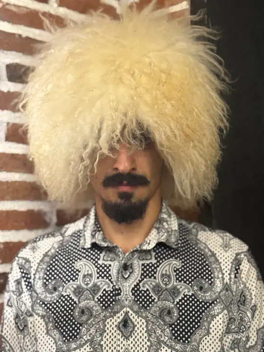 White sheepskin papakha with long wavy wool, worn by a man in a patterned shirt, front view, brick wall behind