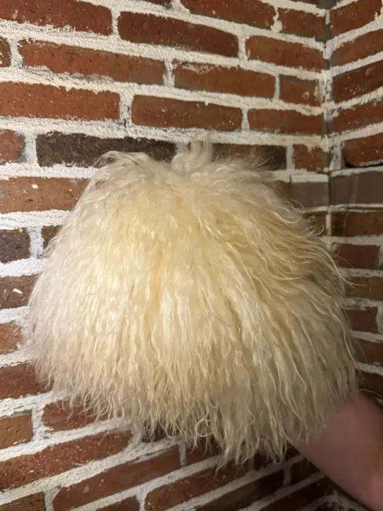 White sheepskin papakha held in a hand against a brick wall, seen from the side