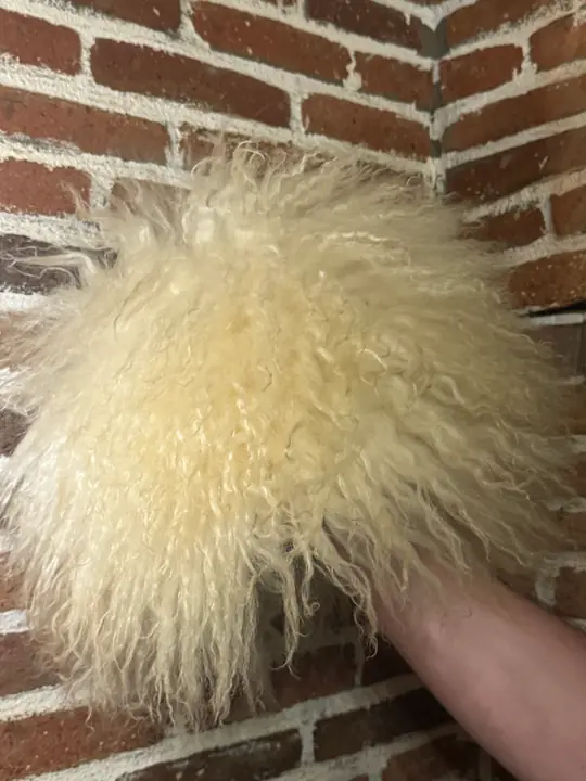 White sheepskin papakha held in a hand against a brick wall, showing the long wool