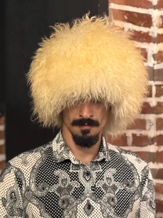 White sheepskin papakha with long wavy wool, worn by a man in a patterned shirt, front view, brick wall behind