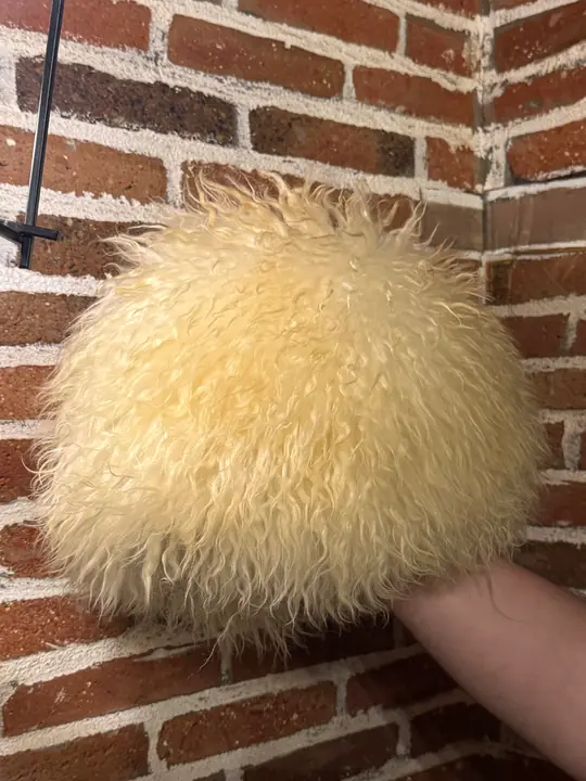 White sheepskin papakha held in a hand against a brick wall, showing the wool